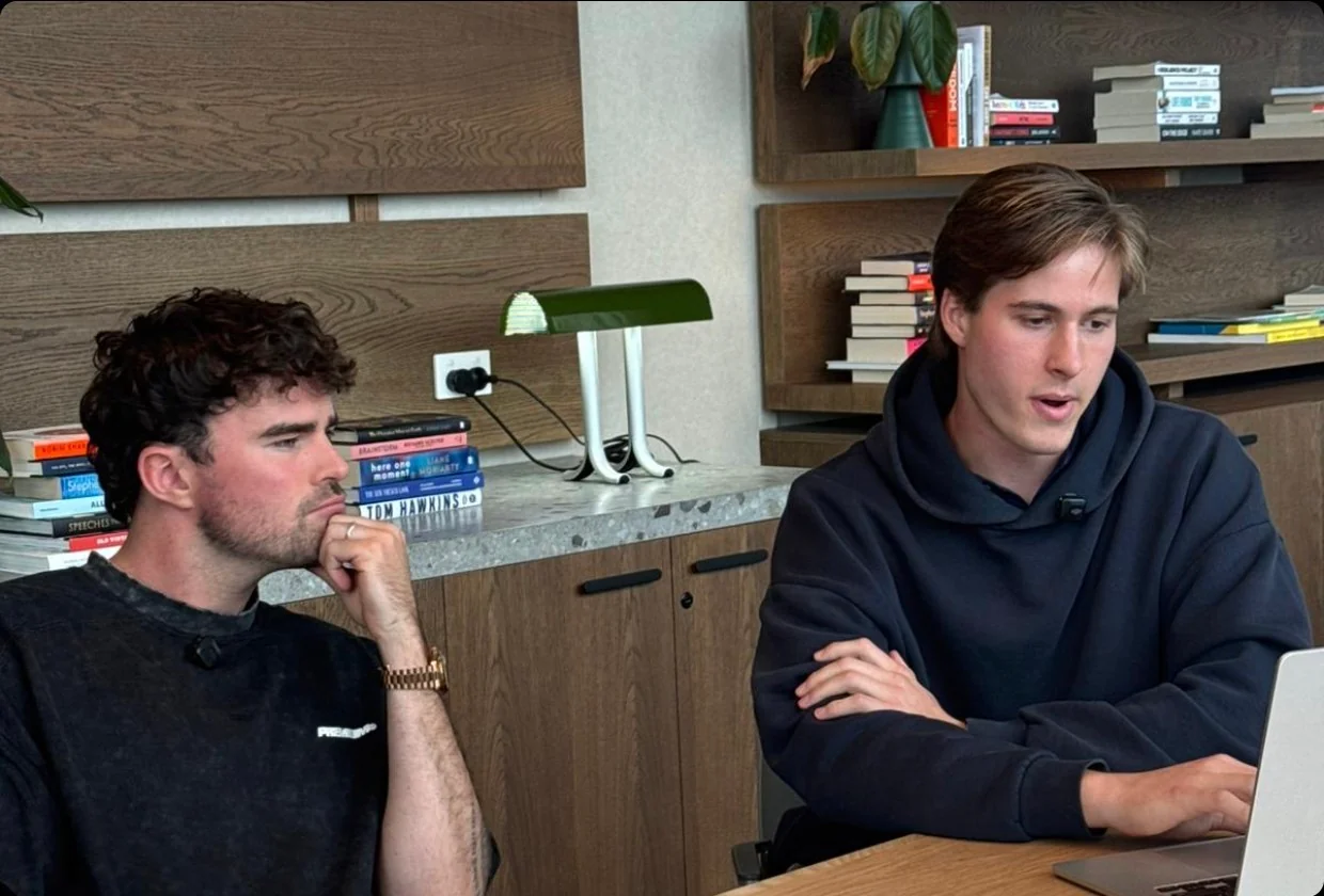 Two founders at a desk, one working through a task on a laptop while the other thinks it over